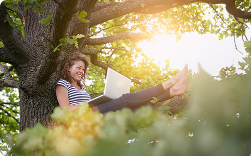 Frau sitzt mit Laptop am Baum und lächelt